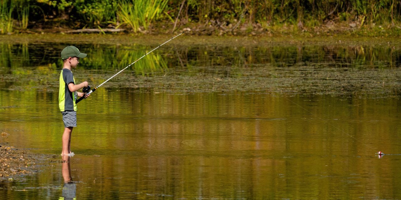 A Cub Scout fishing from the bank, with the rod reflected in the water